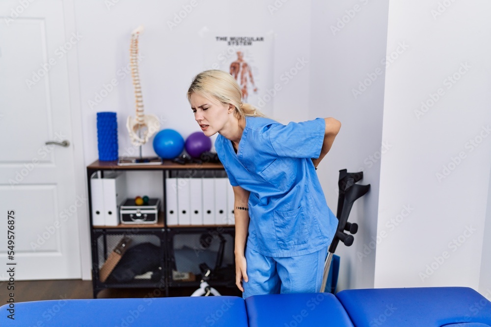 Poster Young physiotherapist woman working at pain recovery clinic suffering of backache, touching back with hand, muscular pain