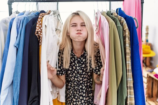Young blonde woman searching clothes on clothing rack puffing cheeks with funny face. mouth inflated with air, catching air.