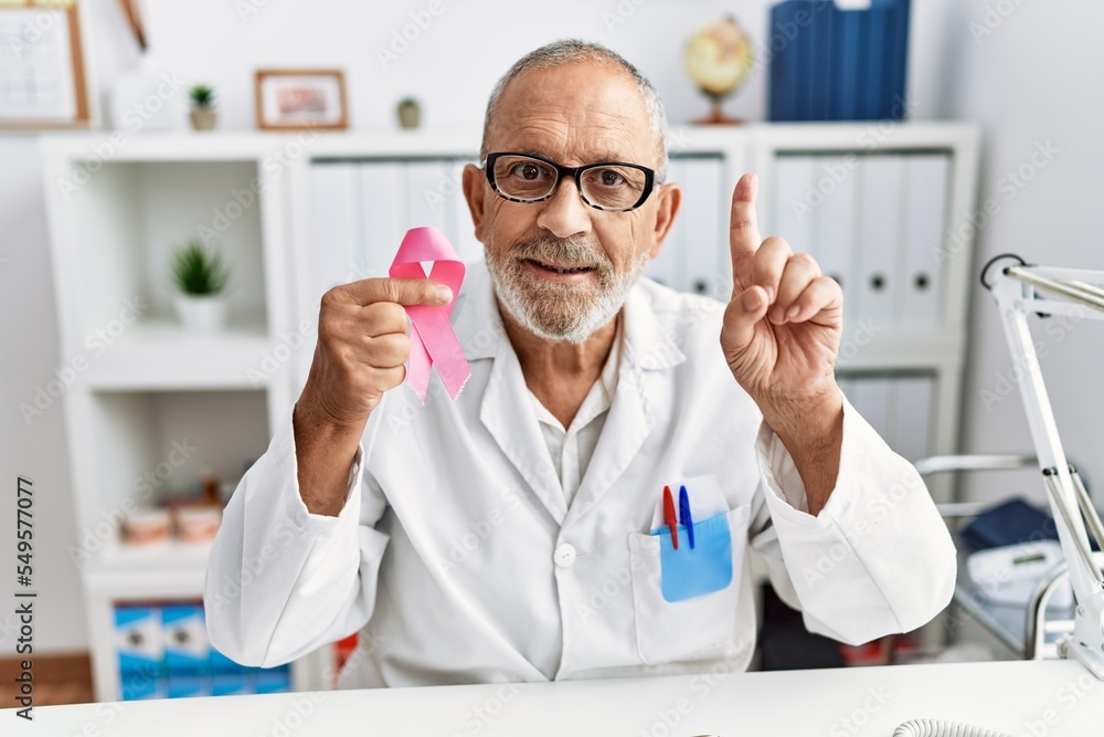 Poster mature doctor man holding pink cancer ribbon at the clinic surprised with an idea or question pointi