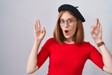 Young redhead woman standing wearing glasses and beret looking surprised and shocked doing ok approval symbol with fingers. crazy expression