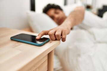 Young hispanic man turning off smartphone alarm lying on the bed at bedroom.