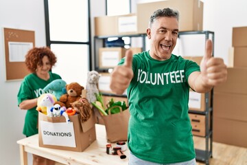 Middle age man wearing volunteer t shirt at donations stand approving doing positive gesture with hand, thumbs up smiling and happy for success. winner gesture.