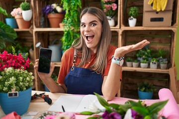 Hispanic young woman working at florist shop showing smartphone screen celebrating victory with...