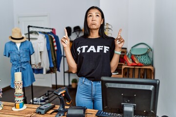 Young hispanic woman working as staff at retail boutique pointing up looking sad and upset,...