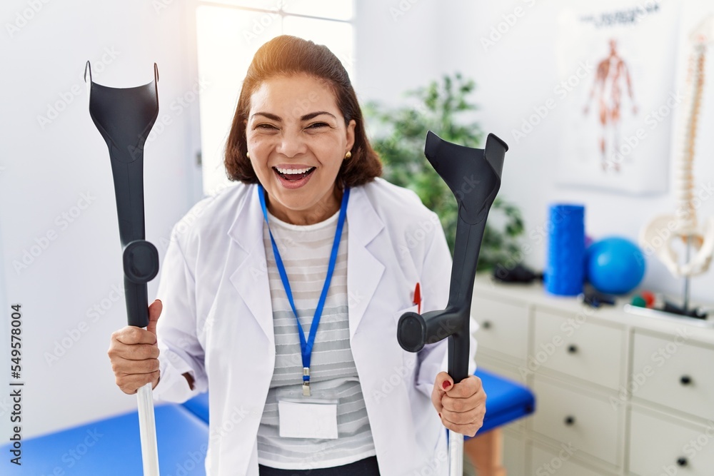 Canvas Prints Middle age hispanic physiotherapy woman holding crutches smiling and laughing hard out loud because funny crazy joke.