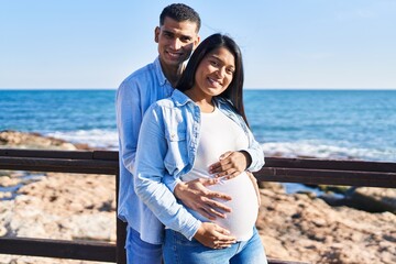 Young latin couple expecting baby hugging each other standing at seaside