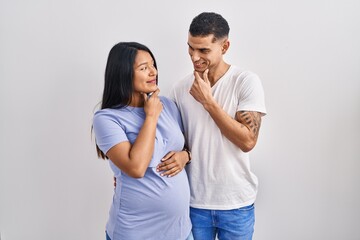 Fototapeta premium Young hispanic couple expecting a baby standing over background looking confident at the camera smiling with crossed arms and hand raised on chin. thinking positive.
