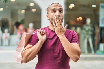 Hispanic man with beard holding shopping bags outdoors covering mouth with hand, shocked and afraid...