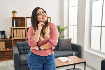 Young hispanic woman working at the office wearing glasses smiling looking confident at the camera with crossed arms and hand on chin. thinking positive.