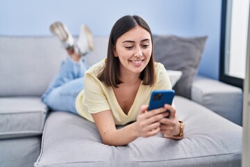 Young hispanic woman using smartphone lying on sofa at home