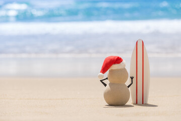 Sandy Christmas Snowman is watching the waves, standing on beautiful beach with a surf board -...