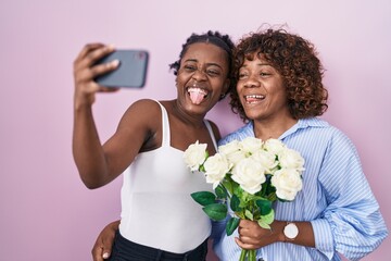 Two african women taking a selfie photo with flowers sticking tongue out happy with funny expression.