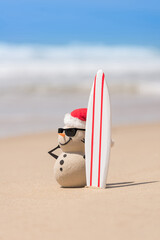 Sandy Christmas Snowman is watching the waves, standing on beautiful beach with a surf board