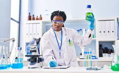 Fototapeta premium African american woman scientist holding bottle writing on document at laboratory