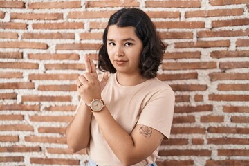 Fototapeta na wymiar Young hispanic woman standing over bricks wall holding symbolic gun with hand gesture, playing killing shooting weapons, angry face