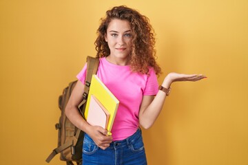 Young caucasian woman wearing student backpack and holding books smiling cheerful presenting and pointing with palm of hand looking at the camera.