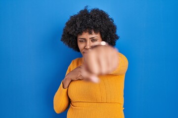 Fototapeta na wymiar Black woman with curly hair standing over blue background punching fist to fight, aggressive and angry attack, threat and violence
