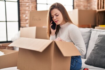 Young blonde woman unboxing package talking on the smartphone at new home