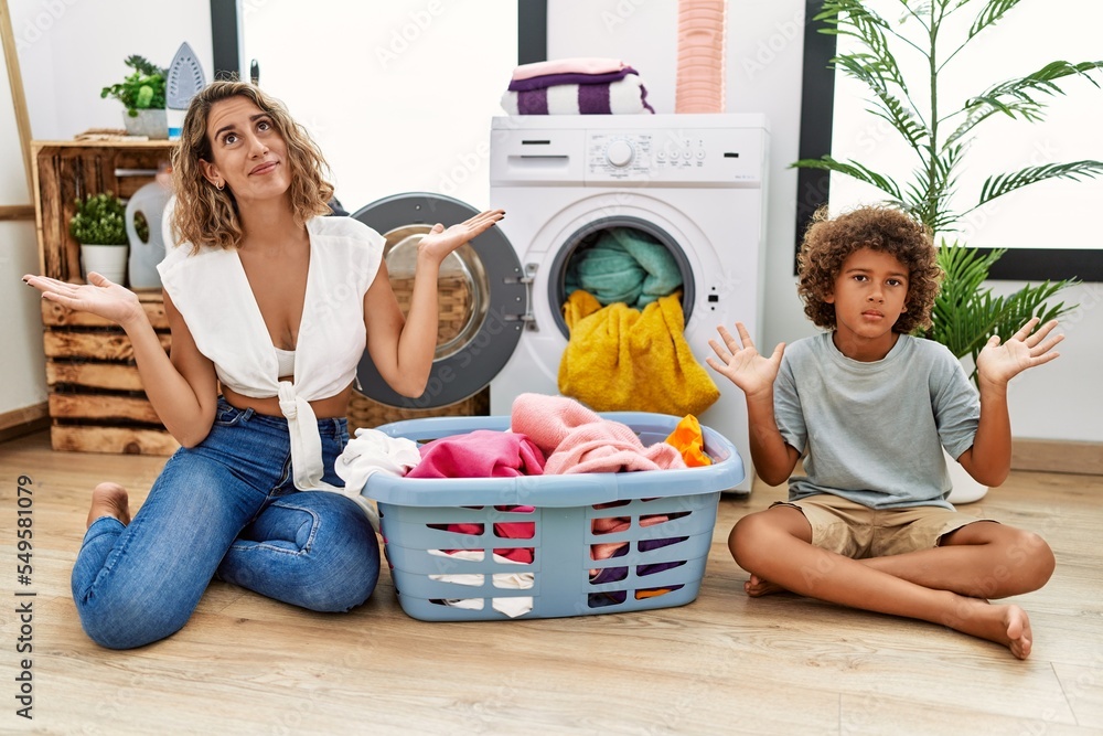 Sticker Young woman and son putting dirty laundry into washing machine clueless and confused expression with arms and hands raised. doubt concept.