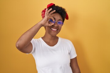Young african american woman standing over yellow background doing ok gesture with hand smiling, eye looking through fingers with happy face.