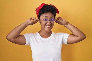 Young african american woman standing over yellow background smiling pulling ears with fingers,...