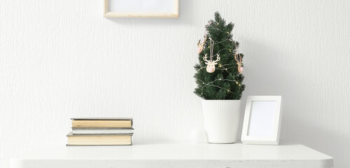 Small Christmas tree with photo frame and books on table near light wall