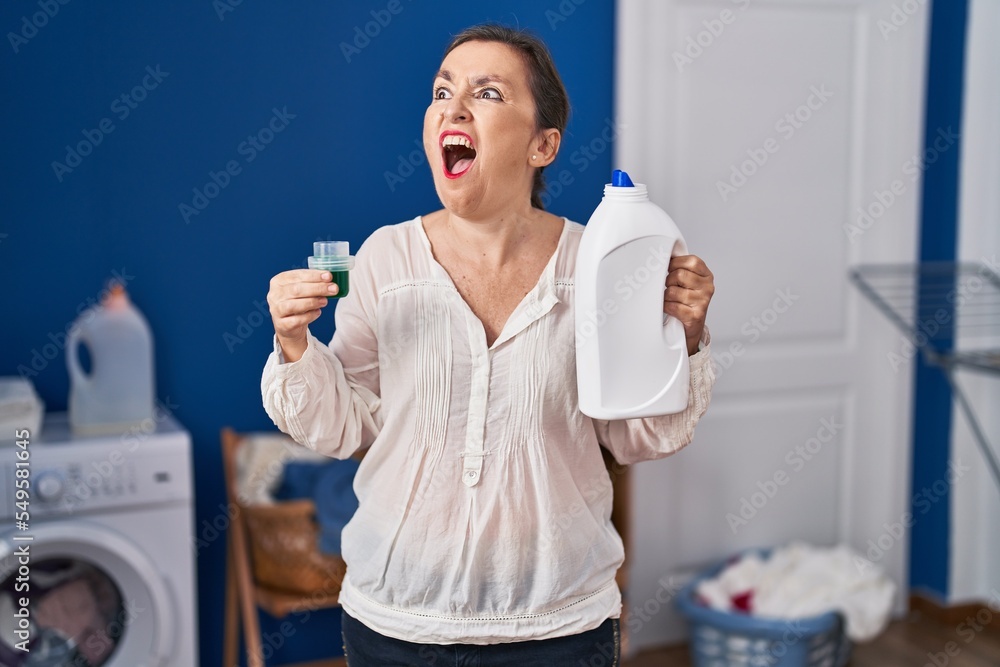 Wall mural Middle age hispanic woman holding detergent bottle angry and mad screaming frustrated and furious, shouting with anger. rage and aggressive concept.