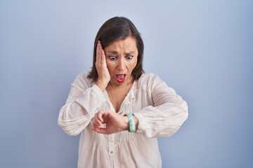 Middle age hispanic woman standing over blue background looking at the watch time worried, afraid...