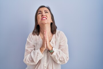 Middle age hispanic woman standing over blue background begging and praying with hands together...