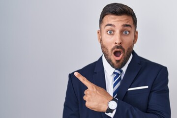 Handsome hispanic man wearing suit and tie surprised pointing with finger to the side, open mouth...