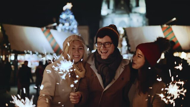 Happy multiracial friends waving bengal firework sparklers at Christmas eve. People celebrate New year at decorated background outdoors at night party smile and hug. Winter holiday or vacation concept
