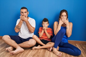 Family of three sitting on the floor at home shocked covering mouth with hands for mistake. secret...