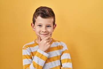 Young caucasian kid standing over yellow background looking confident at the camera with smile with crossed arms and hand raised on chin. thinking positive.