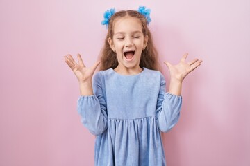 Young little girl standing over pink background celebrating mad and crazy for success with arms...