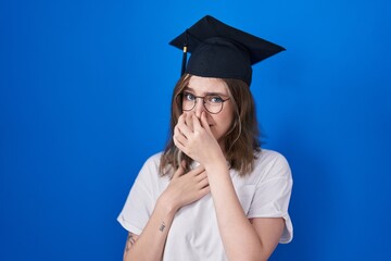 Blonde caucasian woman wearing graduation cap smelling something stinky and disgusting, intolerable smell, holding breath with fingers on nose. bad smell