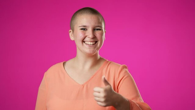 Pretty gender fluid non binary young woman 20s with hearing aid turns around look at camera showing thumbs up like gesture portrait isolated on pink background in studio.