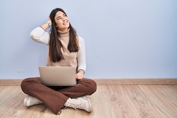 Young brunette woman working using computer laptop sitting on the floor smiling confident touching hair with hand up gesture, posing attractive and fashionable