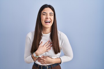 Young brunette woman standing over blue background smiling and laughing hard out loud because funny...
