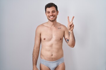 Fototapeta premium Young hispanic man standing shirtless wearing underware showing and pointing up with fingers number two while smiling confident and happy.