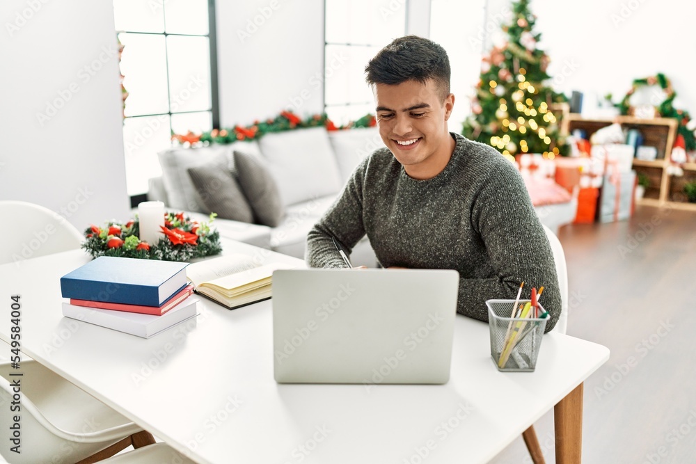 Sticker Young hispanic man studying using laptop sitting by christmas tree at home