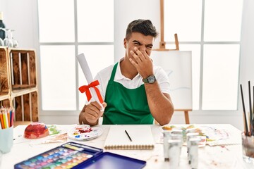 Young hispanic man at art studio holding degree smelling something stinky and disgusting,...