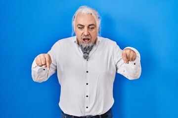 Fototapeta premium Middle age man with grey hair standing over blue background pointing down with fingers showing advertisement, surprised face and open mouth