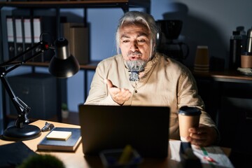 Middle age man with grey hair working at the office at night looking at the camera blowing a kiss with hand on air being lovely and sexy. love expression.