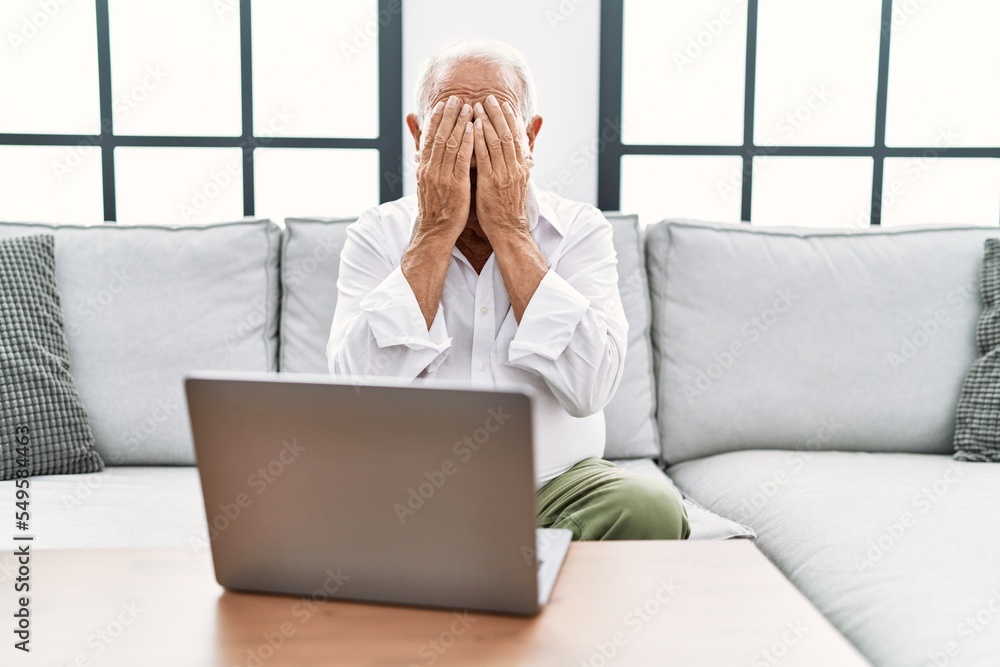 Sticker senior man using laptop at home sitting on the sofa with sad expression covering face with hands whi