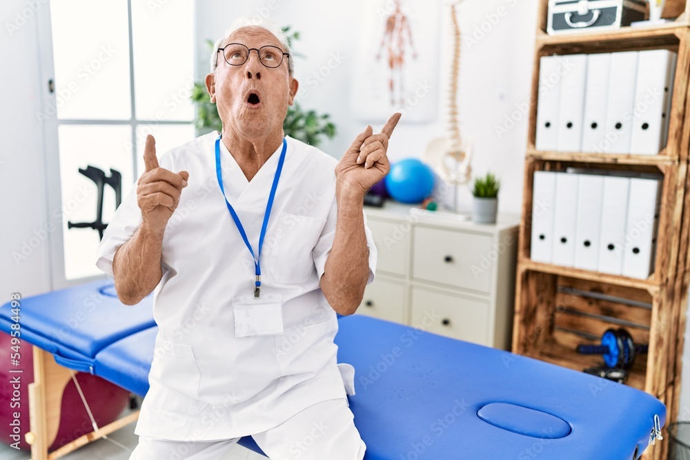 Wall mural senior physiotherapy man working at pain recovery clinic amazed and surprised looking up and pointin