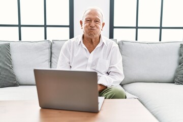 Senior man using laptop at home sitting on the sofa puffing cheeks with funny face. mouth inflated with air, crazy expression.