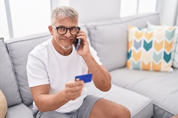 Middle age grey-haired man talking on smartphone and credit card sitting on sofa at home