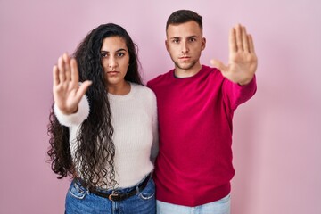 Young hispanic couple standing over pink background doing stop sing with palm of the hand. warning...