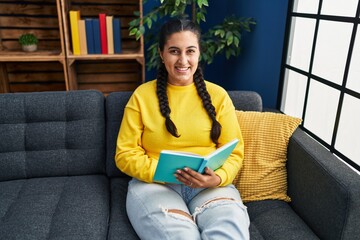 Young hispanic woman reading book sitting on sofa at home