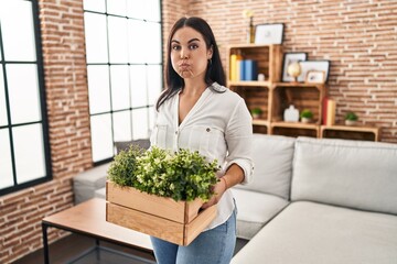 Young hispanic woman holding green plants at the living room puffing cheeks with funny face. mouth inflated with air, catching air.